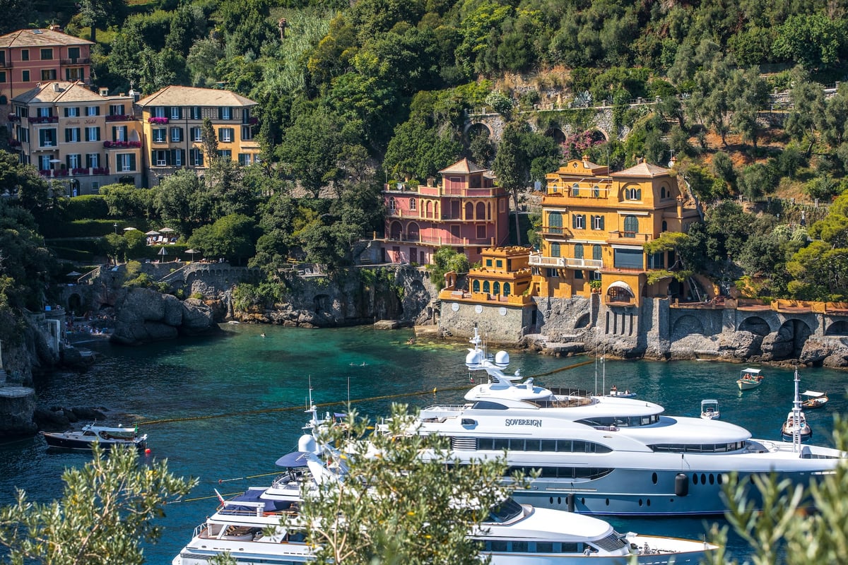 A vibrant view of Portofino, Italy, showing colorful villas perched along the lush green hillside overlooking a turquoise bay filled with luxury yachts.